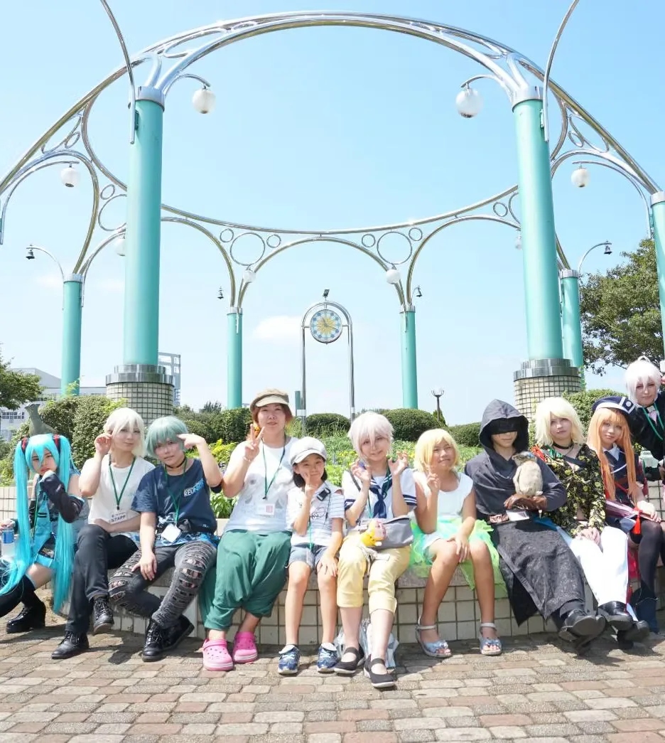 公園の時計塔の前に並んで座るコスプレイヤーとスタッフの集合写真